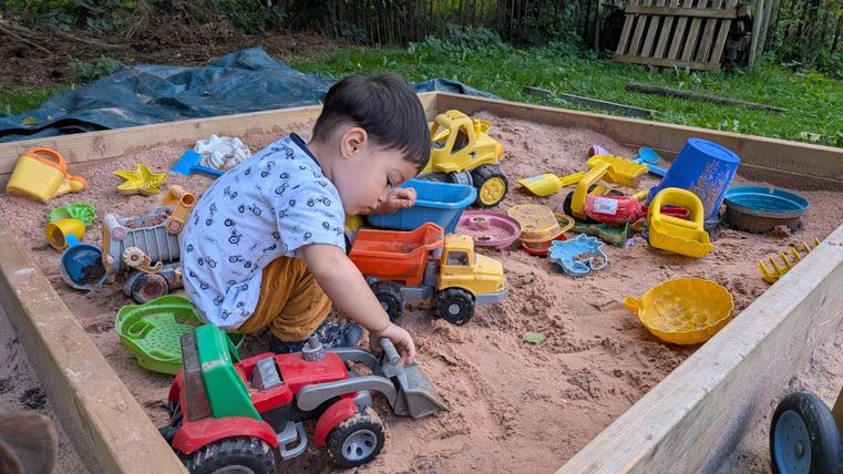 A small child is playing in a sandpit full of colorful toys. It is digging in the sand with a shovel and surrounded by tractors and other construction vehicles.