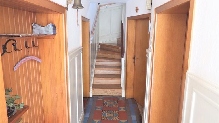 A bright hallway with wooden paneling and a clear staircase in the background. Some coat hooks hang on the walls, and there is a beautiful floor with a colorful pattern.