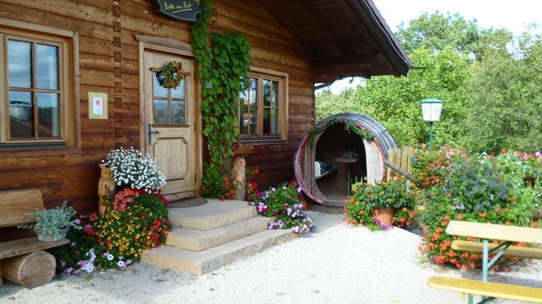 Ein rustikales Holzhaus mit einer einladenden Veranda und blühenden Blumenbeeten. Ein schöner Gartenbereich mit Bänken und einer dekorativen Skulptur ergänzt die ländliche Atmosphäre.