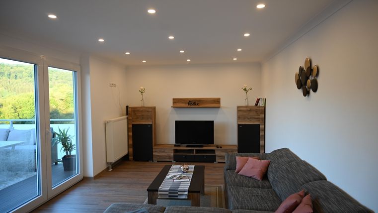 Een moderne woonkamer met een comfortabele bank en een televisie. De ruimte heeft grote ramen die uitzicht op groen bieden.