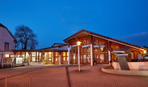 Ein modernes Gebäude mit Holzverkleidung, das bei Dämmerung beleuchtet ist. Es gibt eine einladende Atmosphäre mit Bäumen im Hintergrund.