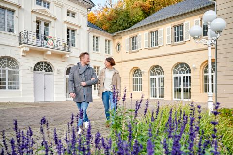 A couple is walking through a picturesque garden with purple flowers. In the background, elegant buildings with large windows can be seen.