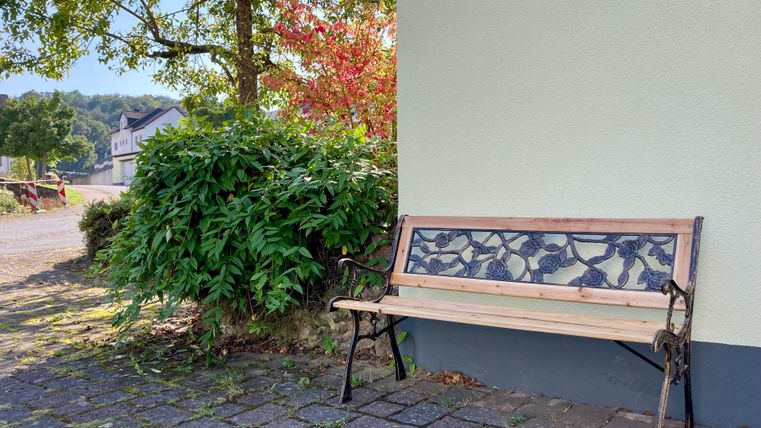 Eine Holzbank steht an einer Wand neben einem grünen Busch. Der Bereich ist ruhig und einladend, umgeben von Bäumen und einem klaren Himmel.