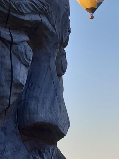 Een grote houten sculptuur van een gezicht staat op de voorgrond. Op de achtergrond zweeft een luchtballon met de tekst "Technis".