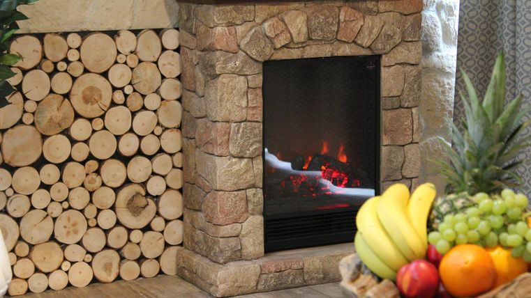 Ein gemütlicher Kamin mit einem Steingehäuse und künstlichen Flammen. Daneben liegt ein Holzstapel und eine Obstschale mit bunten Früchten.