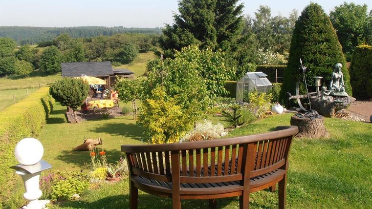 Ein schöner Garten mit einer Holzbank, Bäumen und einem Blick auf die umliegende Landschaft. Im Hintergrund ist eine kleine Hütte sichtbar.
