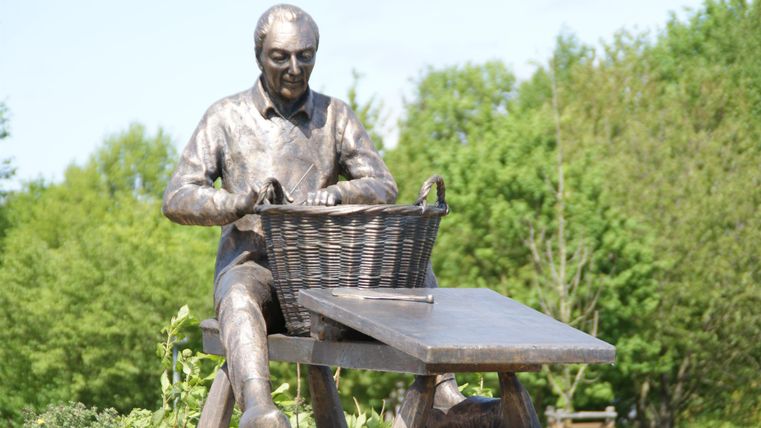 Eine Statue eines Mannes, der an einem Tisch sitzt und einen Korb bearbeitet. Im Hintergrund sind grüne Bäume zu sehen.