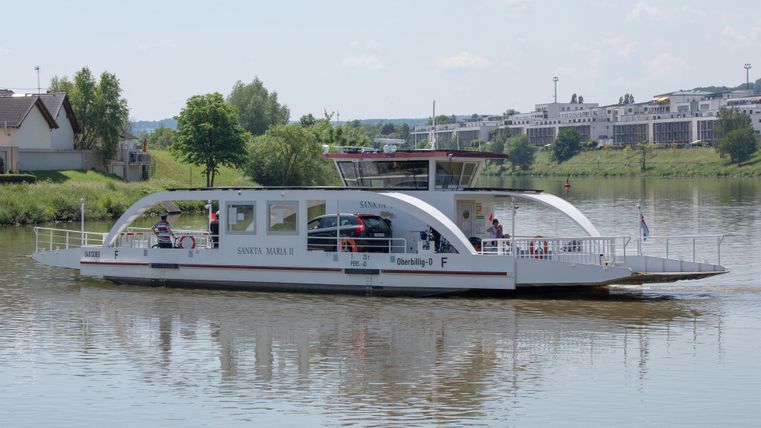 A modern ferry boat is sailing on a calm body of water. Trees and buildings are visible in the background.