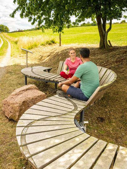 Eine moderne, geschwungene Sitzgelegenheit im Freien. Zwei Personen sitzen zusammen und unterhalten sich in einer grünen Landschaft.