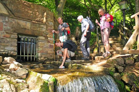 Een groep wandelaars staat bij een kleine waterval in de natuur. Op de achtergrond is een stenen gebouw te zien, omgeven door bomen.