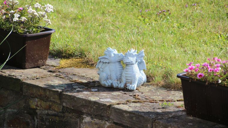 Eine steinerne Drachenfigur steht auf einer Mauer neben blühenden Pflanzen. Im Hintergrund erstreckt sich eine grüne Wiese.