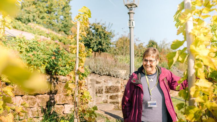 A smiling woman stands in a garden with grapevines. In the background, green plants and a lamp post are visible.