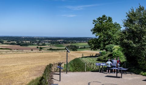 Eine Aussicht von einem Aussichtspunkt auf eine weite Landschaft mit Feldern und Bäumen. Zwei Personen stehen am Aussichtspunkt und betrachten die Umgebung.
