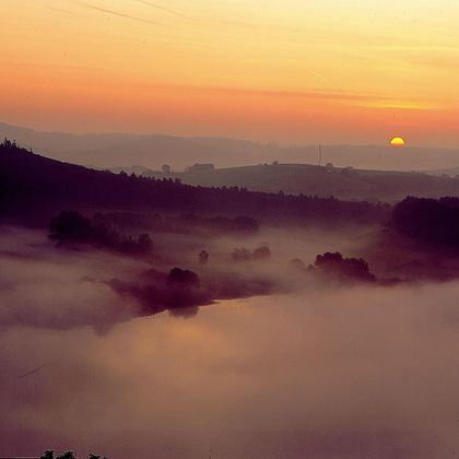 Eine malerische Landschaft bei Sonnenaufgang. Sanfte Hügel und Nebel umgeben einen ruhigen Fluss.