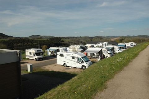 Wohnmobilstellplatz mit mehreren Wohnmobilen, umgeben von grüner Landschaft und Hügeln in der Ferne. Der Himmel ist klar und blau.
