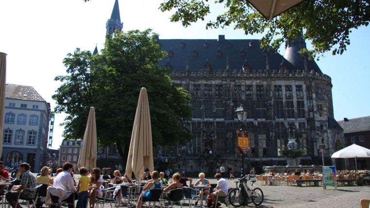 Ein belebter Platz mit vielen Tischen im Freien und Besuchern, die das schöne Wetter genießen. Im Hintergrund ist ein historisches Gebäude zu sehen, umgeben von Bäumen.