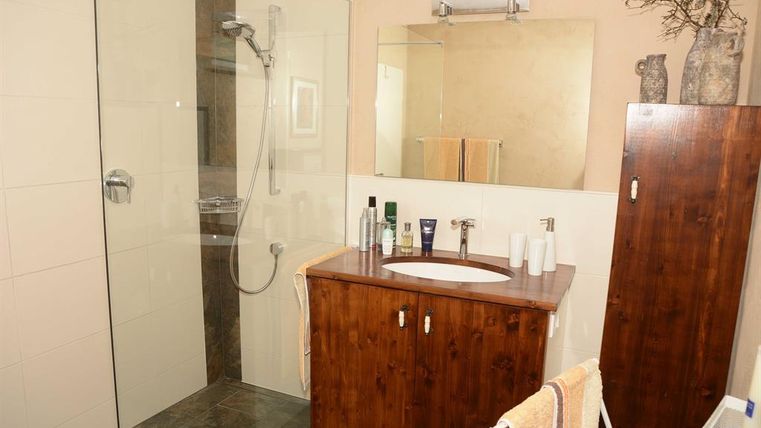 A modern bathroom with a glass shower cabin and a wooden furniture. The mirror reflects little light, and various grooming products are placed on the washbasin.