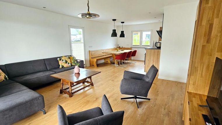 Ein modernes Wohnzimmer mit einem großen grauen Sofa und einem Holztisch. Es gibt auch einen Essbereich mit roten Stühlen und großen Fenstern, die viel Licht hereinlassen.