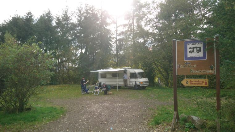 Ein Wohnmobil steht auf einem Campingplatz umgeben von Bäumen. Ein Schild weist auf den Platz hin.