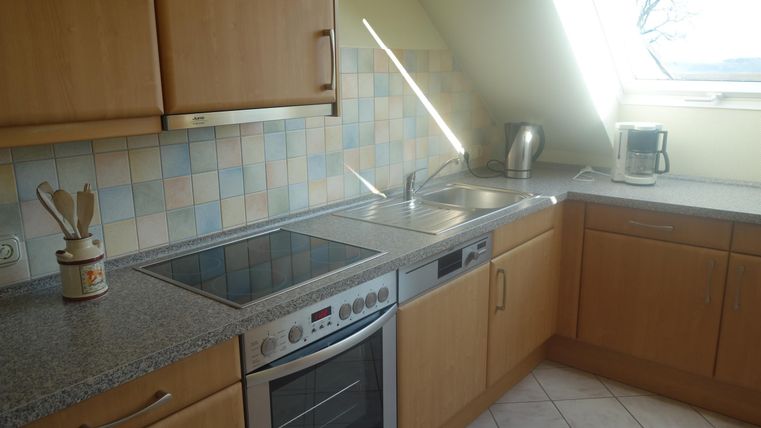 Een moderne keuken met houten kasten en een lichte tegelsvloer. Het werkblad is van graniet en er zijn verschillende keukentoestellen.