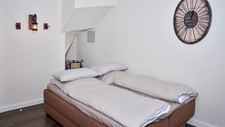 A cozy bedroom with a large bed and a modern wall clock. The walls are painted light and the floor is made of wood.