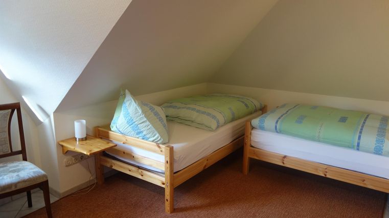 Een eenvoudige slaapkamer met twee eenpersoonsbedden en een stoel. De muren zijn licht en de vloeren zijn met tapijt bedekt.