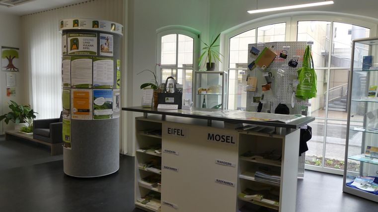 Een moderne receptie met een hoge tafel en informatiestands. Het licht komt binnen via grote ramen en de omgeving lijkt uitnodigend en vriendelijk.