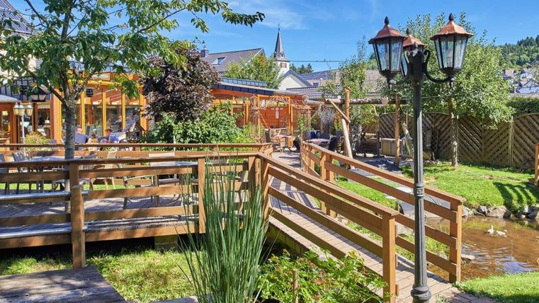 Ein schöner Garten mit Holzbalkonen, Pflanzen und einem kleinen Gewässer. Im Hintergrund sind ein Gebäude und ein Kirchturm sichtbar.