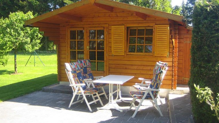 Een gezellig houten huis met een klein terras en twee tuinstoelen. De tuin is groen en helder verlicht door de zon.