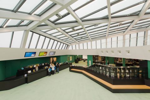 A modern reception area with large windows and bright light. People are standing at a reception desk, and there are shelves in the background.