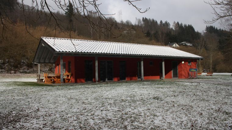 Ein rotes Gebäude mit einem weißen Dach steht in einem schneebedeckten Feld. Im Hintergrund sind Bäume und sanfte Hügel sichtbar.