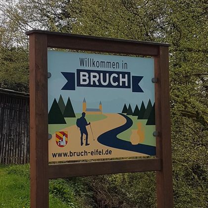 Een welkomstbord voor de gemeente Bruch. Het bord toont een landschap met een wandelaar en een weg.