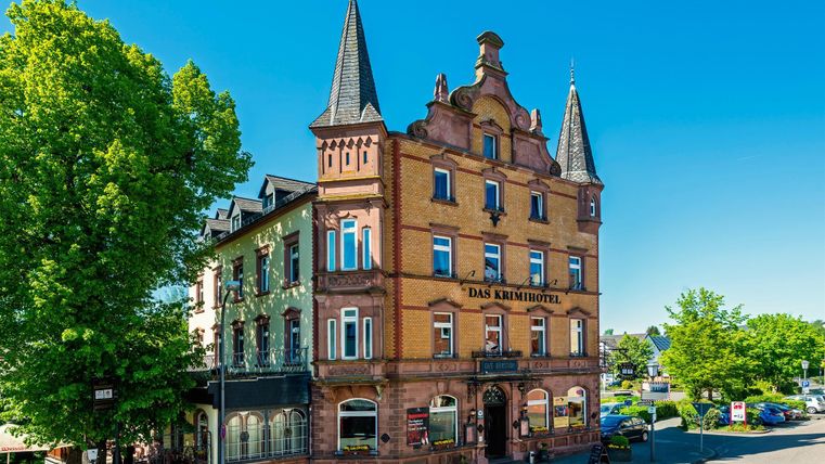 Historisch gebouw met torens en de inscription 'Het Krimihotel', omringd door bomen en een blauwe hemel.