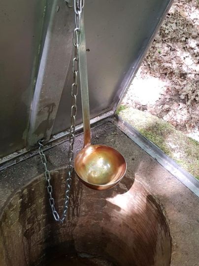 Ein offener Brunnen mit einem Eimer aus Metall, der an einer Kette hängt. Der Brunnen ist von Natur umgeben und hat trübes Wasser.