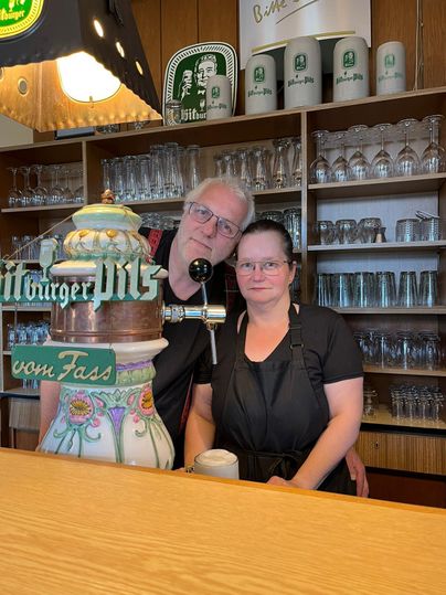 A bartender couple stands behind the bar in a pub. They smile warmly and are surrounded by glasses and beer mugs.
