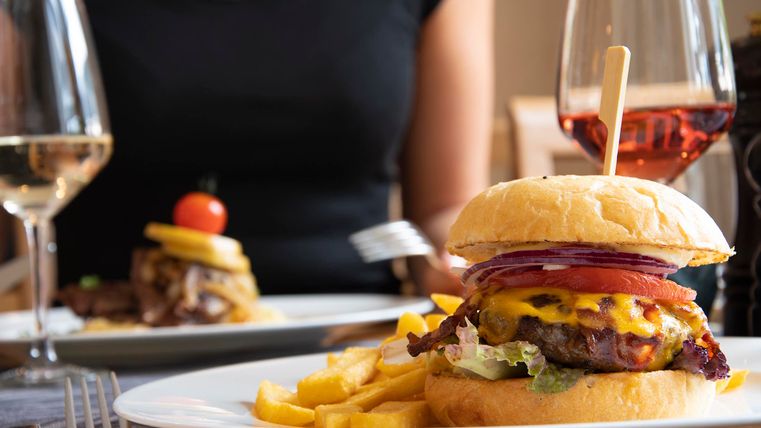 A delicious hamburger with cheese, fresh vegetables, and French fries. In the background, other dishes and drinks can be seen.