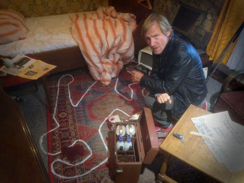 Ein Mann kniet in einer Dachkammer neben einer Kreidezeichnung eines Körpers auf einem Teppich. Ein Arztkoffer und Papiere liegen auf einem Tisch.