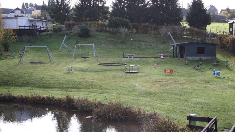 Ein Spielplatz mit Schaukeln, Rutschen und einem kleinen Teich. Im Hintergrund sind Bäume und ein Spielhaus zu sehen.