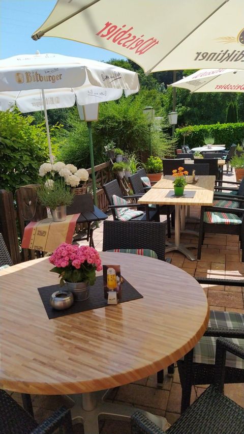 Ein einladender Außenbereich mit Tischen und Stühlen, umgeben von grünen Pflanzen. Auf den Tischen stehen Blumen und Sonnenschirme spenden Schatten.