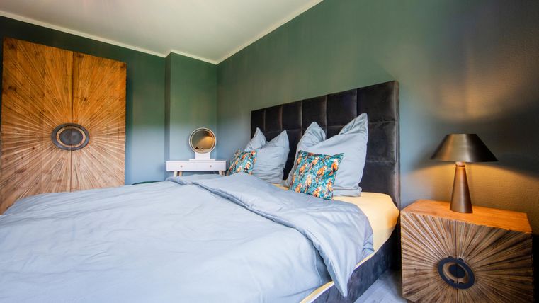 Ein modernes Schlafzimmer mit einem bequemen Bett und stilvollen Kissen. Die Wände sind in einem beruhigenden Grünton gestrichen, und es gibt eine Holzkommode sowie eine elegante Tischlampe.
