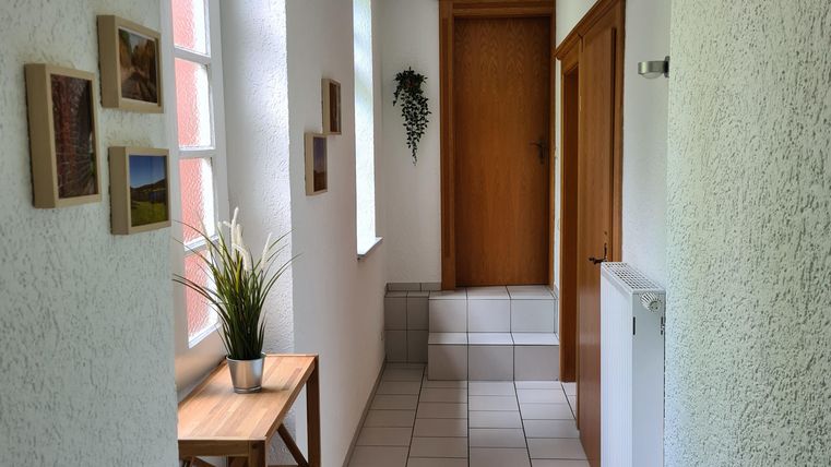 Ein heller Flur mit weißen Wänden und Fliesenboden. Eine kleine Holzkommode und einige Bilder schmücken den Raum.