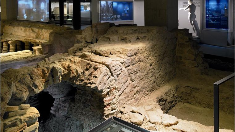 Eine archäologische Ausgrabung in einem Museum mit sichtbaren Steinen und Überresten. Im Hintergrund sind digitale Ausstellungen zu sehen.