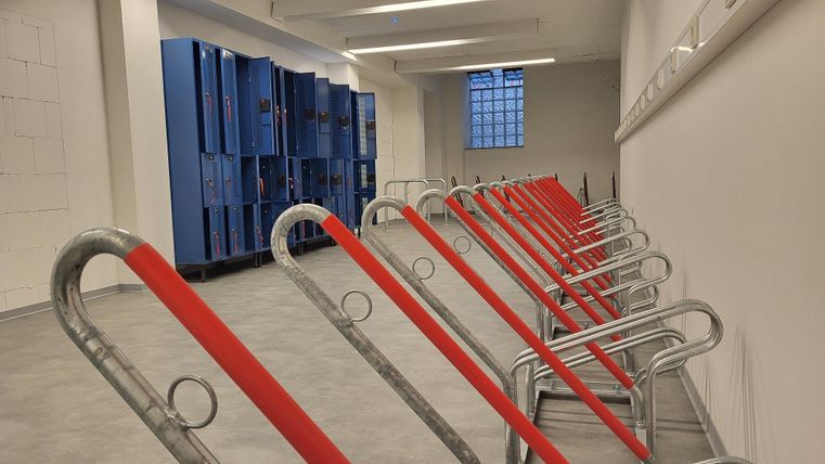 Een lege ruimte met blauwe kasten aan de muur. Voor staan meerdere metalen afzettingen met rode linten.