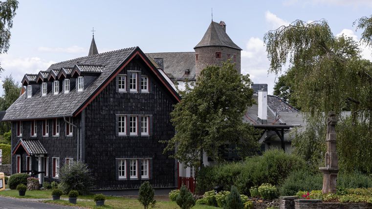 Ein charmantes Gebäude mit dunklen Schindeln und einem Turm. Es ist von üppigem Grün umgeben und hat eine einladende Atmosphäre.