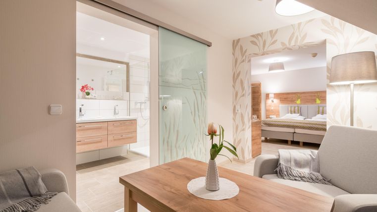 Ein modernes Zimmer mit einem eleganten Tisch und einem gemütlichen Sessel. Im Hintergrund sind ein Badezimmer und ein Schlafzimmer zu sehen, die durch eine Glastür getrennt sind.