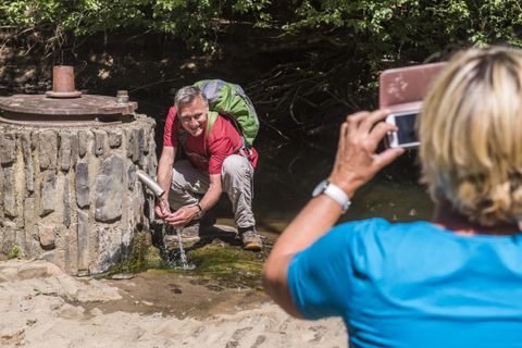 Ein Mann füllt eine Flasche mit Wasser aus einer Quelle. Eine Frau fotografiert ihn lächelnd von hinten.