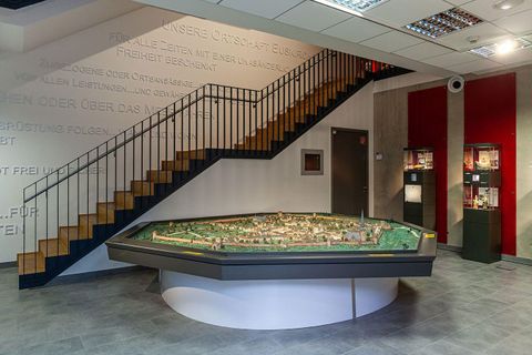 Ein Raum mit einer detaillierten Stadtmodellierung in der Mitte. Eine Treppe führt nach oben, während die Wände mit Texten geschmückt sind.