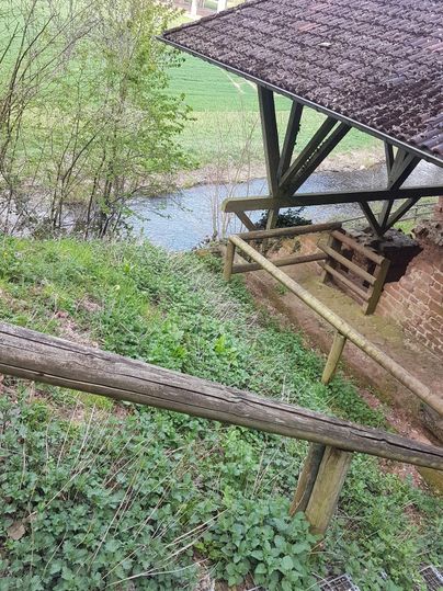 Ein ruhiger Fluss fließt unter einem Holzbau. Umgeben von Grün und Bäumen vermittelt die Szenerie eine friedliche Atmosphäre.