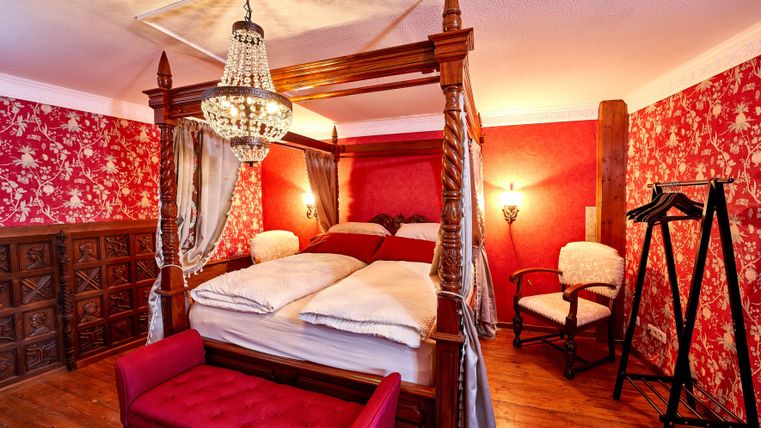 An elegant bedroom with a four-poster bed and red wallpaper. The room is cozy furnished and exudes a warm atmosphere.