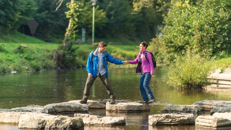 Zwei Personen überqueren Trittsteine in einem Fluss.